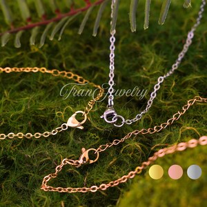 May include: Close-up of three delicate necklaces in gold, silver, and rose gold. Each chain features a lobster clasp closure. The background is a bed of green moss and foliage. The text "Tran Jewelry" is visible.