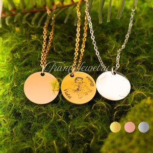 May include: Three round pendant necklaces with chains. One is gold-toned, one is silver-toned, and one is gold-toned with an engraved design. The engraved pendant has a cartoon character and the names "Violet" and "Elaine". The necklaces are displayed on green moss.