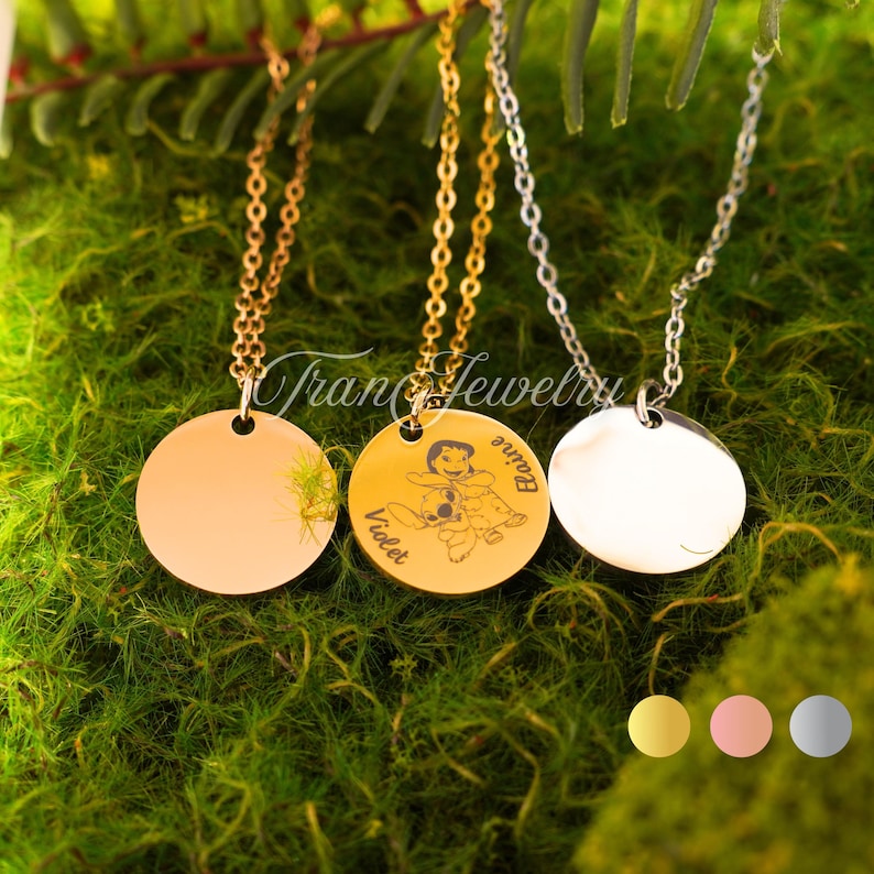 May include: Three round pendant necklaces with chains. One is gold-toned, one is silver-toned, and one is gold-toned with an engraved design. The engraved pendant has a cartoon character and the names "Violet" and "Elaine". The necklaces are displayed on green moss.