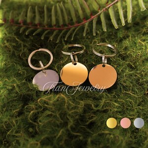 May include: Three metal pet tags in gold, rose gold, and silver, each with a circular pendant and a keyring. The tags are displayed on a bed of green moss with fern accents. The brand name "Fran Jewelry" is visible.