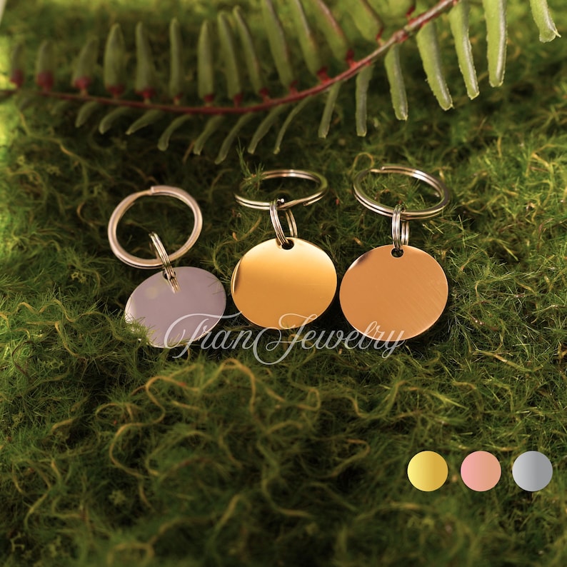 May include: Three metal pet tags in gold, rose gold, and silver, each with a circular pendant and a keyring. The tags are displayed on a bed of green moss with fern accents. The brand name "Fran Jewelry" is visible.