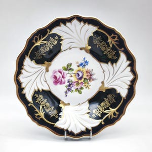 May include: A decorative plate with a black and gold scalloped edge. The plate features a white centre with a floral bouquet of pink, yellow, and purple flowers. The plate has white leaf-like designs and gold accents.