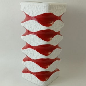 May include: A white ceramic vase with a red and white wavy pattern. The vase is tall and has a square base.