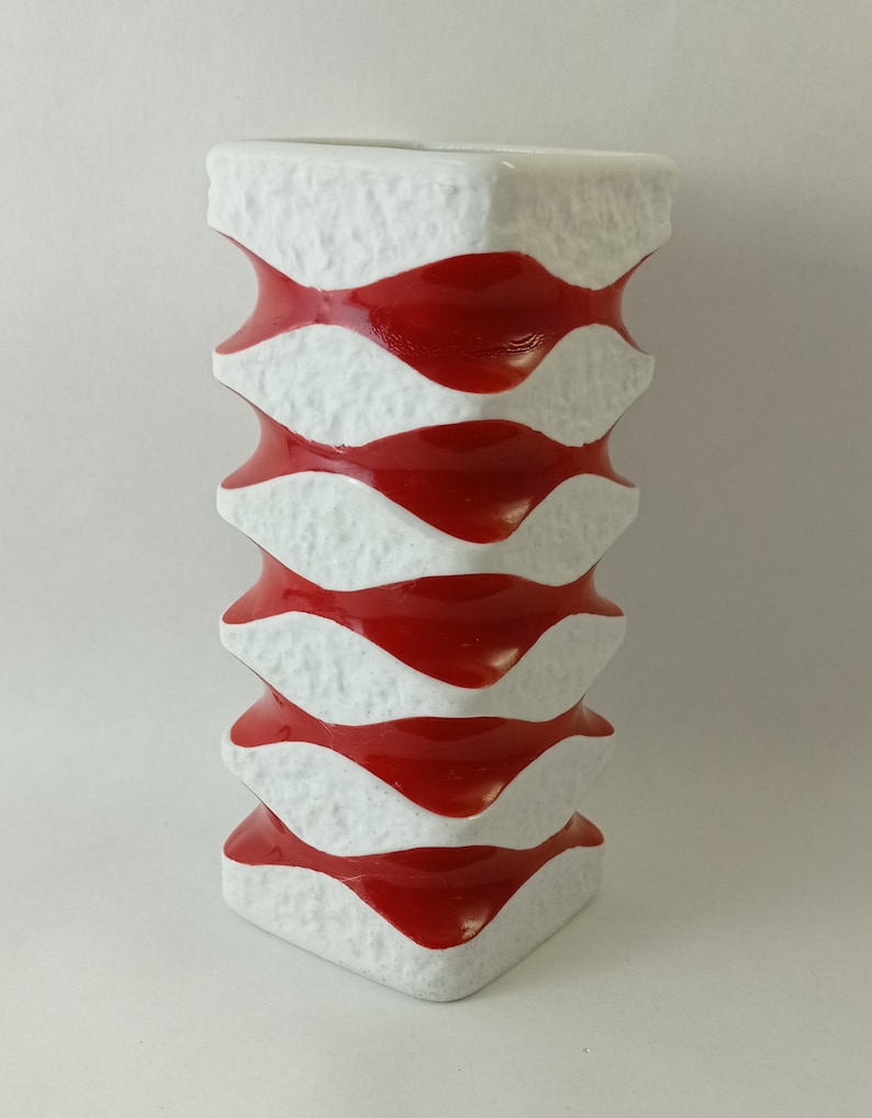 May include: A white ceramic vase with a red and white wavy pattern. The vase is tall and has a square base.