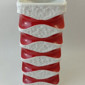May include: A white ceramic vase with a textured surface and red bands. The vase is square in shape and has a modern design.