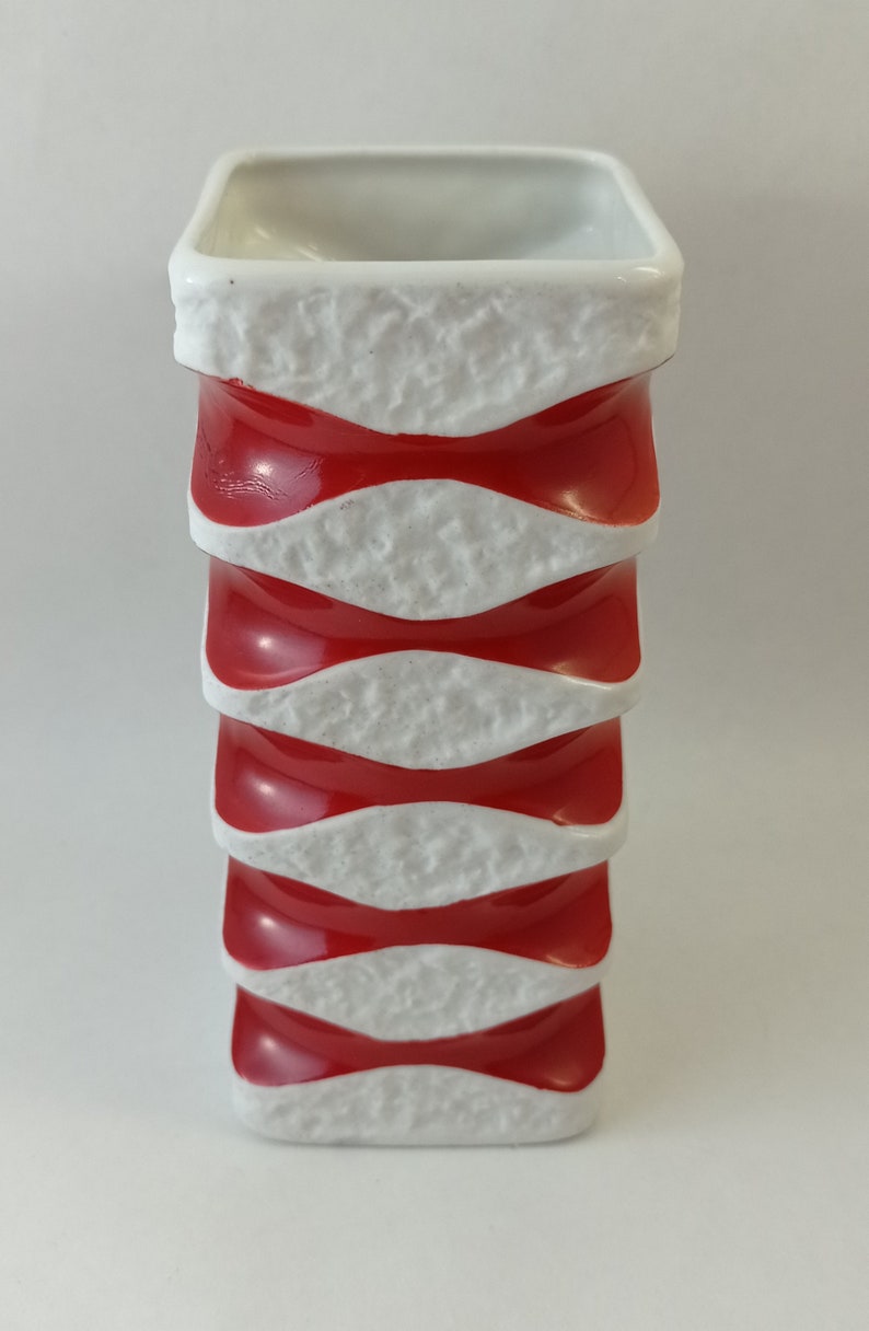May include: A white ceramic vase with a textured surface and red bands. The vase is square in shape and has a modern design.