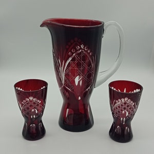 May include: A ruby red glass jug with a clear handle and two matching glasses. The glassware features intricate etched designs. The jug is approximately 23 cm tall, and the glasses are about 10 cm tall.