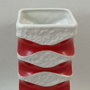 May include: A white ceramic vase with red and white wavy stripes. The vase has a square shape and a textured surface.