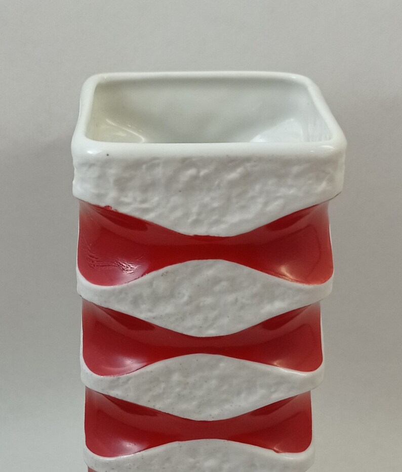May include: A white ceramic vase with red and white wavy stripes. The vase has a square shape and a textured surface.