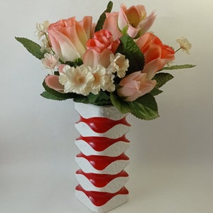 May include: A white ceramic vase with red wavy stripes holds a bouquet of pink and peach roses and white flowers.