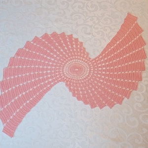 Op de afbeelding: Een roze gehaakt tafellaken met een spiraalvormig ontwerp. Het tafellaken is gemaakt met een delicate, kant-achtige steek en heeft een rond midden.