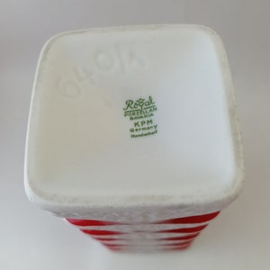 May include: A white porcelain vase with red and white stripes. The base of the vase has a green label that reads "Royal Porzellan Bavaria KPM Germany Handarbeit".