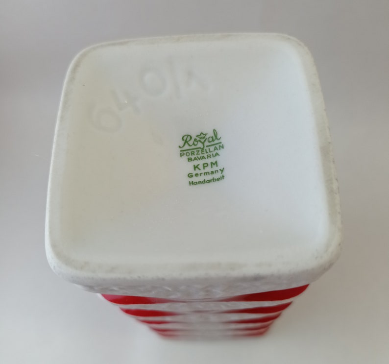 May include: A white porcelain vase with red and white stripes. The base of the vase has a green label that reads "Royal Porzellan Bavaria KPM Germany Handarbeit".