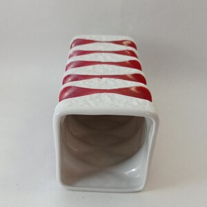 May include: White ceramic vase with red and white stripes. The vase is rectangular and has a square opening at the top.