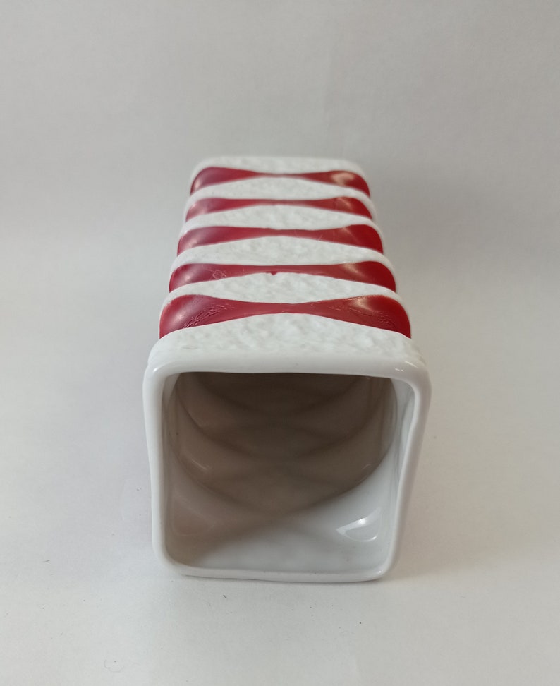 May include: White ceramic vase with red and white stripes. The vase is rectangular and has a square opening at the top.