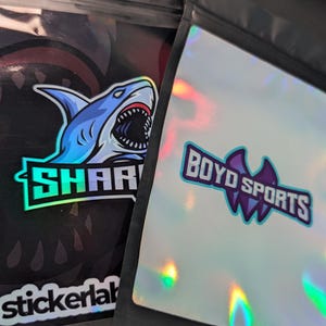 May include: Two holographic mylar bags with zipper closures. One bag has a blue and green shark logo with the text "SHARK" and "stickerlab". The other bag has a purple bat logo with the text "BOYD SPORTS".