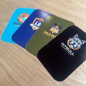 May include: Four different sports card trading cards. The cards are rectangular and feature different logos and designs. The first card is blue with a beach scene and the text "Paradise". The second card is green with a logo that says "Wild Ducks Sports Cards". The third card is black with a logo that says "Pitbull Sports Cards". The fourth card is green with a logo that says "LO Sports Cards".