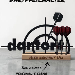 Könnte beinhalten: Ein schwarzer Dartpfeilhalter mit dem Wort "dart" ausgeschnitten. Der Halter hat einen Holzsockel mit dem Text "HIER GEWINNT ULI". Drei Darts mit roten und schwarzen Flights werden angezeigt. Der Text "INDIVIDUELL PERSONALISIERBAR" ist ebenfalls vorhanden.