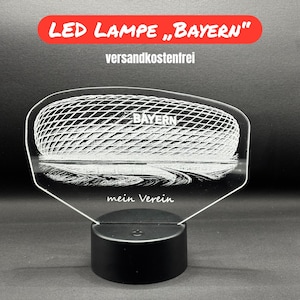 Könnte beinhalten: LED-Lampe mit dem Text "BAYERN" in einem roten Banner. Die Lampe zeigt eine detaillierte Illustration eines Stadions mit dem Wort "BAYERN" und dem Satz "mein Verein". Die Lampe steht auf einem schwarzen Sockel.