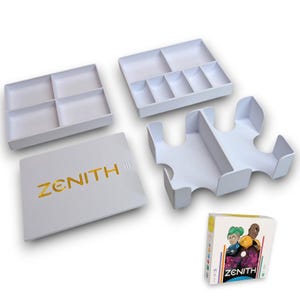 Pode incluir: Conjunto de caixas organizadoras brancas para o jogo de tabuleiro Zenith. O conjunto inclui uma caixa com tampa com o nome do jogo em letras douradas, uma bandeja com seis compartimentos, uma bandeja com três compartimentos e um divisor com quatro compartimentos. A caixa do jogo também é mostrada com uma ilustração colorida dos personagens do jogo.