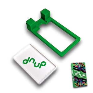 Può includere: Una cornice rettangolare in plastica verde con due blocchi quadrati rialzati a un'estremità, una carta rettangolare bianca con il testo verde "dnup" al centro e una piccola scatola da gioco rettangolare con il testo "dnup" e grafiche colorate.