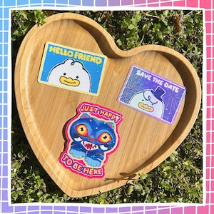 May include: A heart-shaped wooden tray holds three colorful stickers. One says "HELLO FRIEND" with a cartoon duck. Another says "SAVE THE DATE" with a duck in a top hat. The third sticker features a blue cat with the text "JUST HAPPY TO BE HERE".