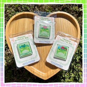 May include: Three acrylic pins featuring a green frog design, each in a clear plastic package. The pins have different phrases: "Overwhelmed" and "Overstimulated". The pins are on a heart-shaped wooden tray.