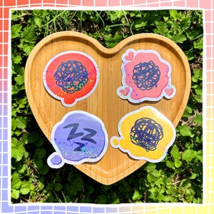 May include: Four die-cut stickers arranged on a heart-shaped wooden tray. The stickers feature colorful speech bubbles in red, pink, purple, and yellow. Three bubbles contain dark blue scribbles, while the purple bubble displays the text ZZZ.
