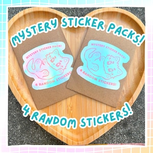 May include: Two brown paper packs with holographic stickers, each labeled "Mystery Sticker Pack!" and "4 Random Stickers!" The packs are on a heart-shaped wooden tray, with the words "Mystery Sticker Packs!" and "4 Random Stickers!" in a playful font.