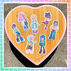 May include: Seven character stickers with a holographic, glittery finish, displayed on a heart-shaped wooden tray with a pink edge. The stickers depict diverse characters in vibrant attire. The initials "PH" are present.