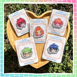 Puede incluir: Cinco pines acrílicos con retratos de personajes de estilo anime, cada uno en una funda de plástico transparente. Los pines tienen pelo rojo, naranja, verde y azul. El embalaje lleva la inscripción "TWST ACRYLIC PIN" y el nombre del artista.