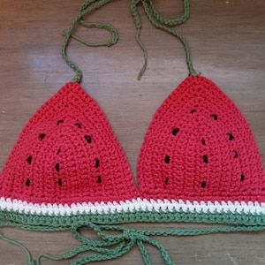 May include: A crocheted bikini top in the shape of a watermelon. The top is red with black seeds and a green top. The bottom of the top is white and green.