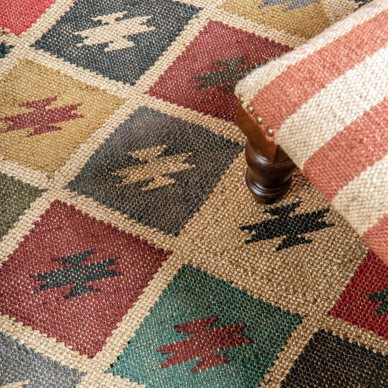 K&ouml;nnte beinhalten: Gewebter Teppich mit einem Rautenmuster in verschiedenen Farben, darunter Rot, Gr&uuml;n, Gold und Grau. Jede Raute weist ein geometrisches Design auf. Ein gestreifter Hocker mit Holzbeinen ist teilweise sichtbar.
