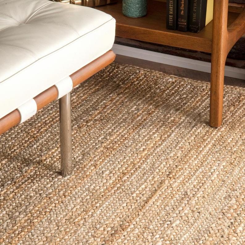 K&ouml;nnte beinhalten: Ein Naturfaserteppich mit geflochtener Textur in Beige- und Braunt&ouml;nen. Der Teppich liegt unter einer wei&szlig;en Lederbank mit Holzakzenten und einem Holzbein. Der Teppich verleiht dem Raum eine rustikale Note.