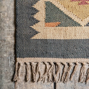 K&ouml;nnte beinhalten: Nahaufnahme eines gewebten Teppichs mit geometrischem Muster. Der Teppich hat einen dunkelgrauen Rand, beige und gr&uuml;ne Dreiecke und einen Fransen aus naturfarbenen Fasern. Der Teppich liegt auf einer Holzoberfl&auml;che.