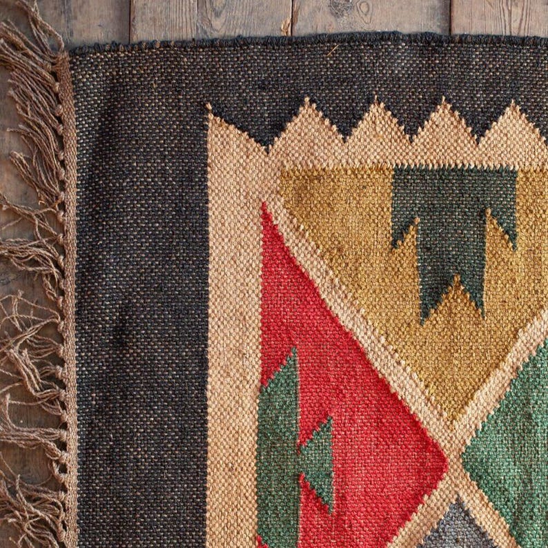 K&ouml;nnte beinhalten: Nahaufnahme eines gewebten Teppichs mit geometrischem Design. Der Teppich hat einen schwarzen Rand, beige, rote, gr&uuml;ne und goldene geometrische Formen. Der Teppich hat eine Fransen an der linken Seite und besteht aus Naturfasern.