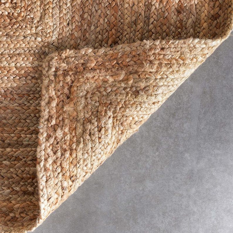 K&ouml;nnte beinhalten: Nahaufnahme eines nat&uuml;rlichen Jute-Teppichs mit geflochtener Textur. Die Ecke des Teppichs ist umgeschlagen und zeigt die Unterseite. Die Farbe ist ein warmes, erdiges Braun mit Farbvariationen. Der Teppich scheint strapazierf&auml;hig und f&uuml;r den Innenbereich geeignet zu sein.