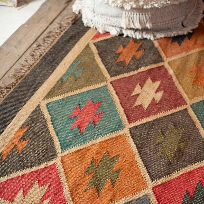 K&ouml;nnte beinhalten: Ein farbenfroher Teppich mit geometrischem Muster. Der Teppich zeigt Quadrate in verschiedenen Farben, darunter Rot, Gr&uuml;n, Orange und Grau, die jeweils ein Sternmuster enthalten. Der Teppich hat einen Fransenrand und besteht aus gewebtem Material.
