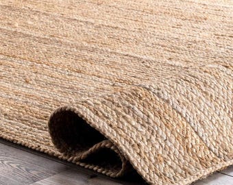 May include: A rectangular area rug crafted from natural tan jute fibers, featuring a tightly woven braided texture. The rug is shown with one corner folded over, revealing the thick, durable construction of the woven plant-based material.