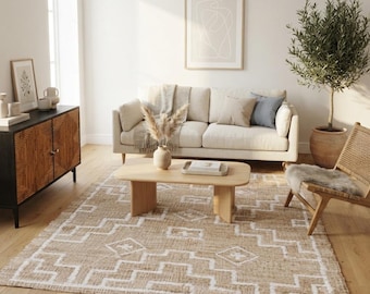 May include: A rectangular area rug featuring a natural tan woven texture with a repeating white geometric pattern of stepped lines and diamond shapes. The rug is positioned on a light wood floor in front of a beige sofa and a wooden coffee table.