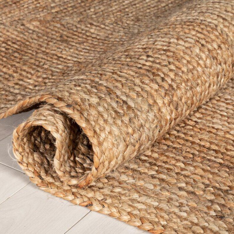K&ouml;nnte beinhalten: Eine aufgerollte Jute-Rolle in Naturt&ouml;nen. Die geflochtene Textur des Teppichs ist sichtbar, mit einem warmen, erdigen Braunton. Das teilweise aufgerollte Design zeigt die Dicke und Konstruktion des Teppichs, was auf einen rustikalen oder b&ouml;hmischen Stil hindeutet.