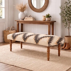 May include: Upholstered bench with a cream-colored fabric featuring a geometric pattern in black, gray, and tan. The bench has turned wooden legs and is placed on a neutral-toned rug. A wooden console table and decorative items are in the background.