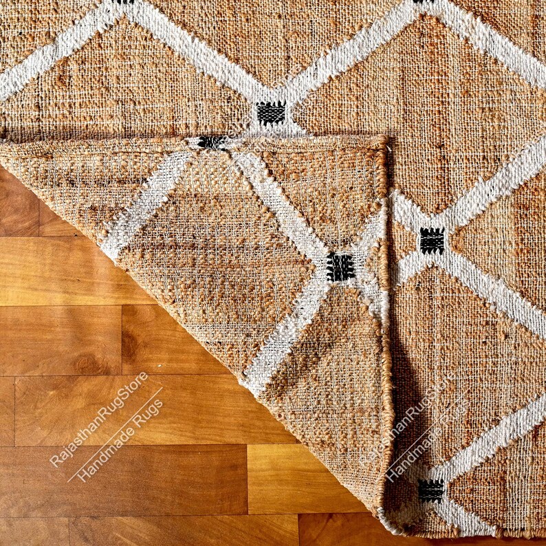 May include: Close-up of a natural fiber rug with a diamond pattern woven in white and black. The rug is folded over, showing the underside of the weave. The text "RajasthaniRugStore Handmade Rugs" is visible in the lower left corner.