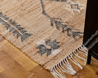 May include: A woven area rug featuring a natural tan jute base with dark charcoal and white geometric patterns. The rug has a textured surface and is finished with long, white fringe tassels along the edge, creating a distinct visual contrast.