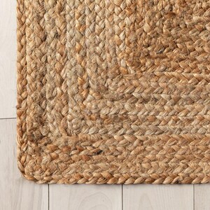 K&ouml;nnte beinhalten: Nahaufnahme eines nat&uuml;rlichen Jute-Teppichs mit geflochtener Textur. Der Teppich hat eine warme, erdige braune Farbe mit Farbvariationen. Die Ecke des Teppichs ist sichtbar und zeigt die detaillierte Webart und Konstruktion. Es ist ein Einrichtungsgegenstand.
