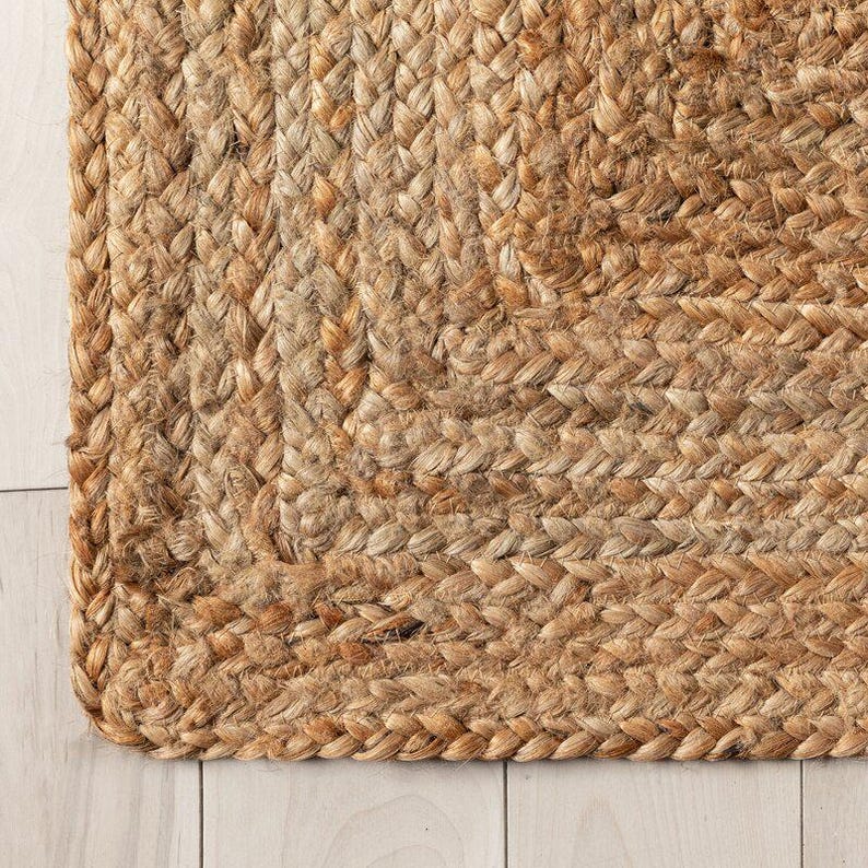 K&ouml;nnte beinhalten: Nahaufnahme eines nat&uuml;rlichen Jute-Teppichs mit geflochtener Textur. Der Teppich hat eine warme, erdige braune Farbe mit Farbvariationen. Die Ecke des Teppichs ist sichtbar und zeigt die detaillierte Webart und Konstruktion. Es ist ein Einrichtungsgegenstand.