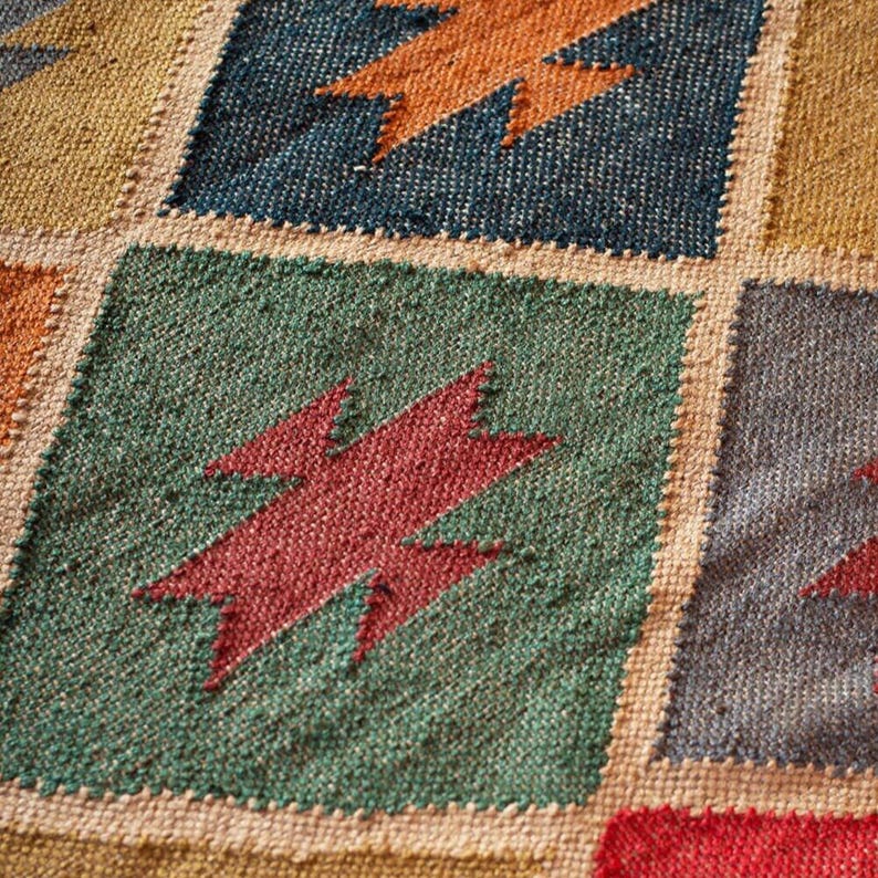 K&ouml;nnte beinhalten: Nahaufnahme eines gewebten Teppichs mit Patchwork-Design. Der Teppich zeigt Quadrate in verschiedenen Farben, darunter Gr&uuml;n, Blau und Gelb, jedes mit einem Blitzdesign in kontrastierenden Farben. Der Teppich hat ein strukturiertes Aussehen.