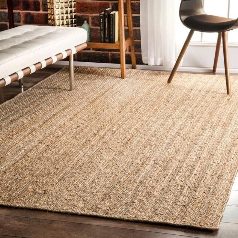 K&ouml;nnte beinhalten: Ein rechteckiger Jute-Teppich in Naturt&ouml;nen mit gewebter Textur. Der Teppich liegt auf einem dunklen Holzboden, und die neutrale Farbpalette erg&auml;nzt die umliegenden M&ouml;bel und die Einrichtung. Der Teppich verleiht dem Raum W&auml;rme und Textur.