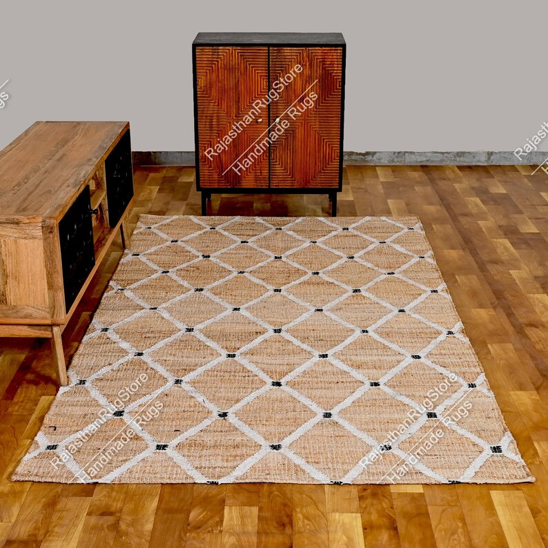 May include: A natural fiber rug with a diamond pattern in white and black on a beige background. The rug is handmade and is shown on a wooden floor with a wooden cabinet in the background.