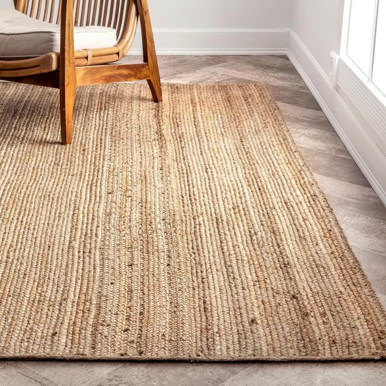 K&ouml;nnte beinhalten: Ein rechteckiger Jute-Teppich in einem nat&uuml;rlichen, hellbraunen Farbton. Der Teppich hat eine gerippte Textur und liegt auf einem gefliesten Boden. Ein Holzstuhl ist teilweise im Hintergrund zu sehen, was auf ein Wohnzimmer oder einen &auml;hnlichen Raum hindeutet.
